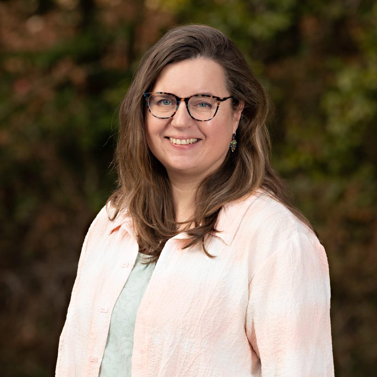 Amy Cooke smiling at the camera wearing glasses, 一件浅蓝绿色衬衫和一件奶油色外套，背景是深绿色和橙色的树叶.