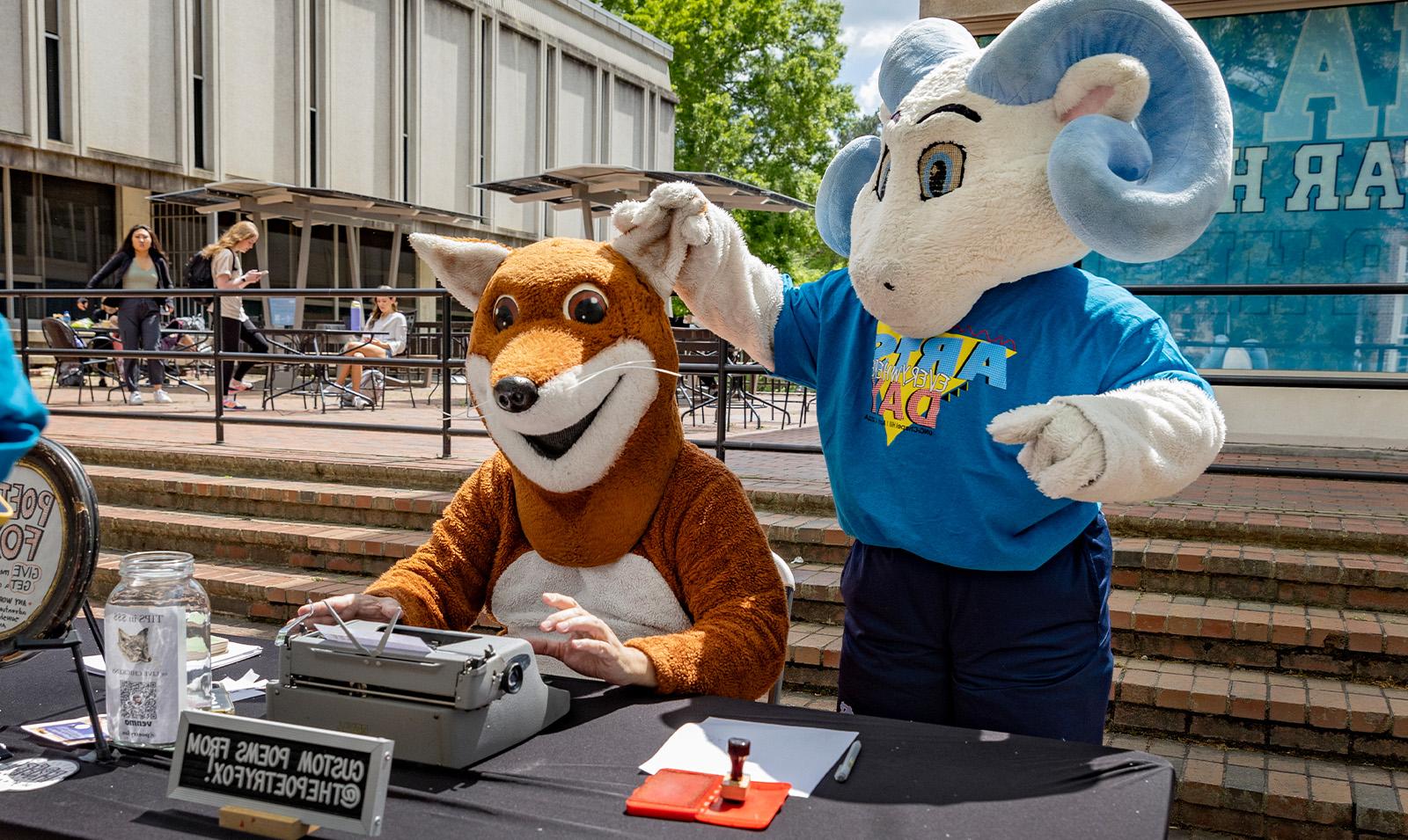 A ram mascot, Rameses Jr., holding the ear of another mascot, Poetry Fox, at Arts Everywhere Day.