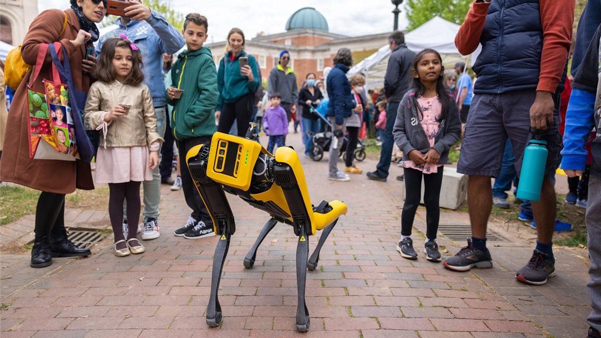 Boston Dynamics four legged robot walking around campus with onlookers of all ages surrounding it.